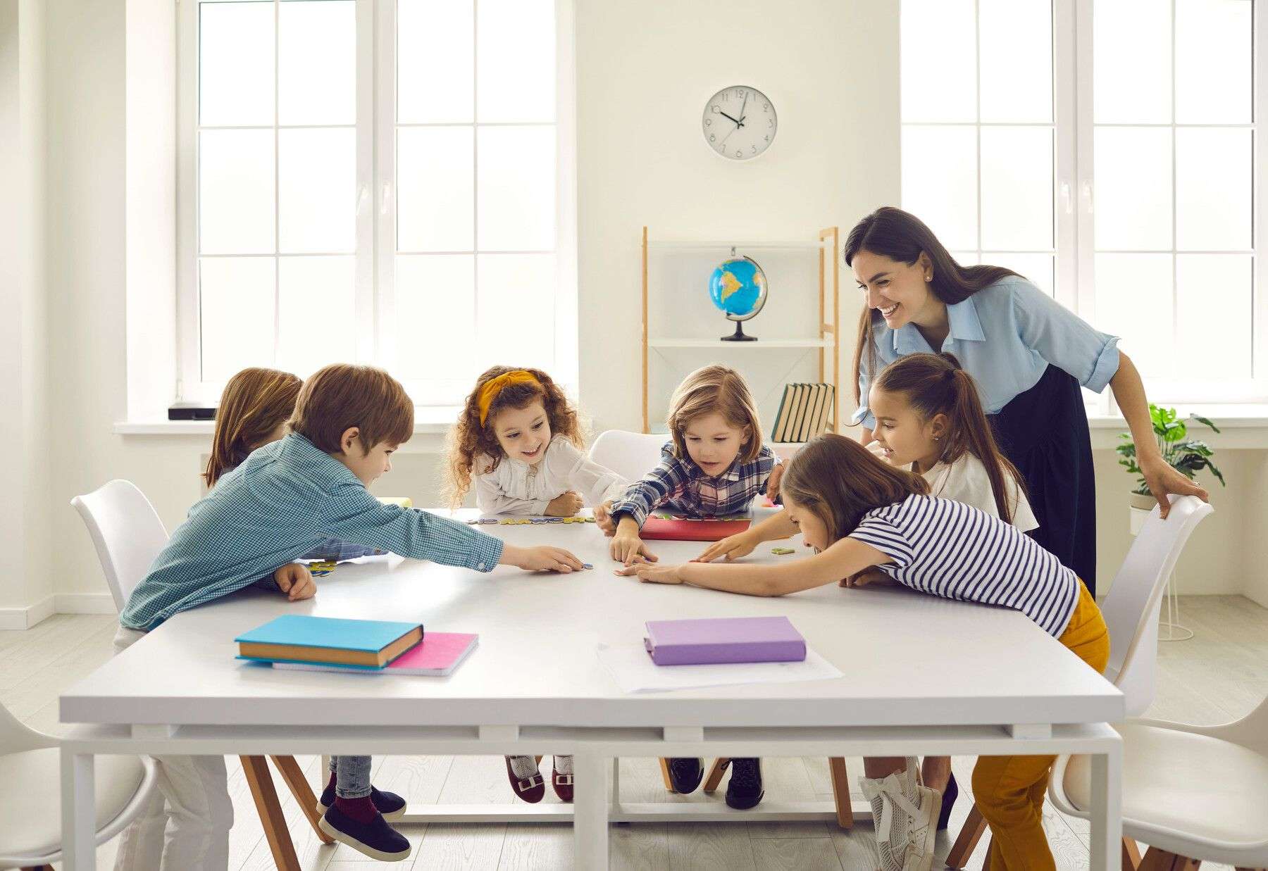 In a bright, airy classroom bathed in natural light from large windows, a cheerful adult woman with dark hair, wearing a light blue blouse and dark skirt, leans over a white table with a warm, encouraging smile, observing a group of five young children intensely engaged in a hands-on activity. The children—three girls and two boys—lean forward with focused expressions, their hands reaching across the table as they manipulate colorful educational toys or puzzle pieces. One girl with curly hair and a yellow headband beams with delight, while another in a striped shirt leans closely, her brow furrowed in concentration. A boy in a blue checkered shirt reaches eagerly toward the center, and another girl in a white shirt and yellow pants is bent low, fully absorbed. The table holds a few brightly colored notebooks—blue, pink, and purple—and papers, suggesting a creative or academic task. Behind them, a small wooden shelf holds a globe and books, and a simple white wall clock indicates the time. The room’s minimalist aesthetic, with white chairs and light wood flooring, enhances the vibrant energy and joyful collaboration among the children and their smiling adult facilitator.