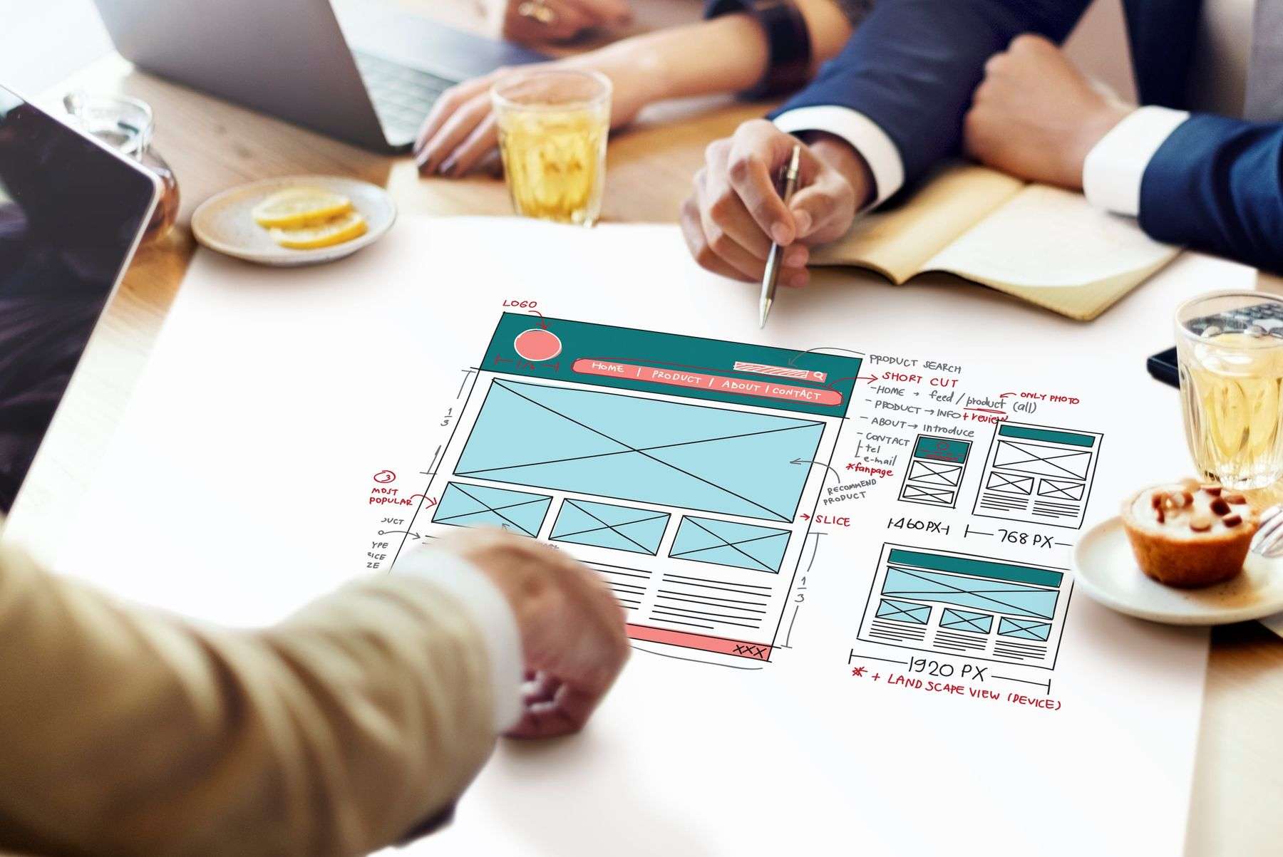 In a warmly lit, modern meeting space, three individuals collaborate around a wooden table, their hands and arms the primary focus as they engage with a hand-drawn website wireframe spread across a large white sheet. One person, wearing a navy suit with a crisp white cuff, leans forward intently, holding a silver pen poised over the design, suggesting active annotation or discussion; their posture conveys focus and involvement. Another participant, clad in a beige blazer, rests their hand near the wireframe, possibly pointing or preparing to contribute. The central design document is vibrant with color-coded sections: a teal header with a red circular logo placeholder, light blue rectangular placeholders for content, and pink and black annotations detailing navigation like “Home,” “Product,” “About,” “Contact,” along with technical specs such as “1460px,” “768px,” and “1920px + LANDSCAPE VIEW (DEVICE).” Surrounding the creative hub are practical elements: a laptop and tablet hint at digital work, a glass of iced tea and a small plate with lemon slices add a touch of refreshment, and a delectable muffin with chocolate sprinkles sits nearby, reinforcing a casual yet productive atmosphere. The scene is one of dynamic, collaborative design thinking, where physical sketches and digital tools converge to shape a digital product.