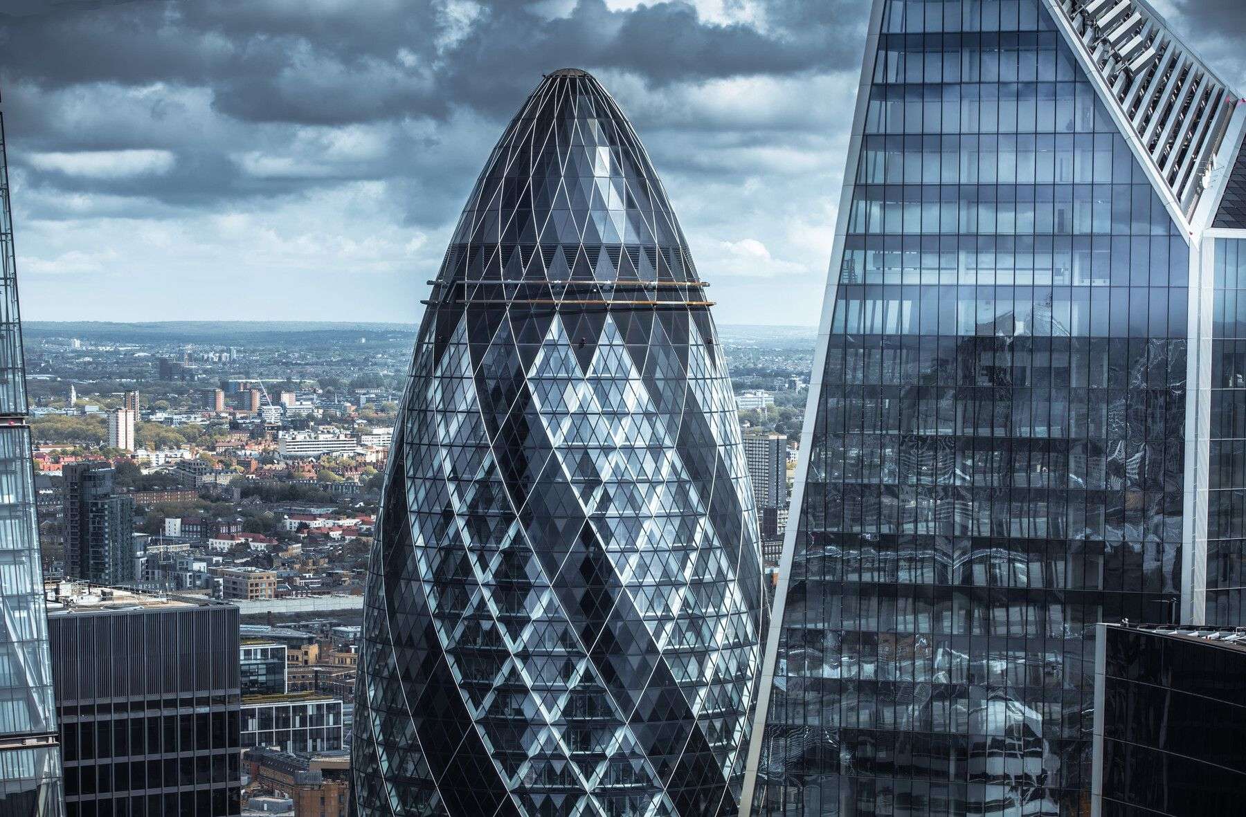 A striking, futuristic skyscraper with a sleek, bulbous, diamond-patterned glass facade dominates the frame, its metallic sheen reflecting the overcast sky, while a sharply angled, glass-clad tower with a white steel truss structure stands beside it, both towering over a sprawling urban landscape beneath a dramatic, cloud-filled sky with hints of pale blue peeking through, casting a moody, cinematic light over the cityscape of rooftops and distant buildings.