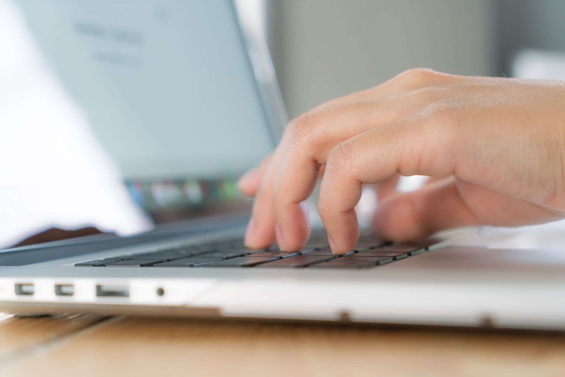 A close-up shot captures a person’s right hand, with fingers delicately positioned over the black keys of a sleek silver laptop, suggesting active typing or navigation; the hand’s skin tone is warm and natural, with subtle texture and faint hair visible on the wrist and knuckles. The laptop, likely a modern MacBook given its slim profile and aluminum finish, rests on a light-brown wooden surface, with its ports—USB and headphone jacks—visible along the side edge. The screen is blurred but emits a soft, cool white glow, hinting at an active display with indistinct icons or text, while the background is intentionally out of focus, featuring muted tones of gray and white that suggest a clean, minimalistic workspace. There’s no visible facial expression or emotion, as the focus is entirely on the tactile interaction between human and machine, emphasizing concentration and digital productivity.