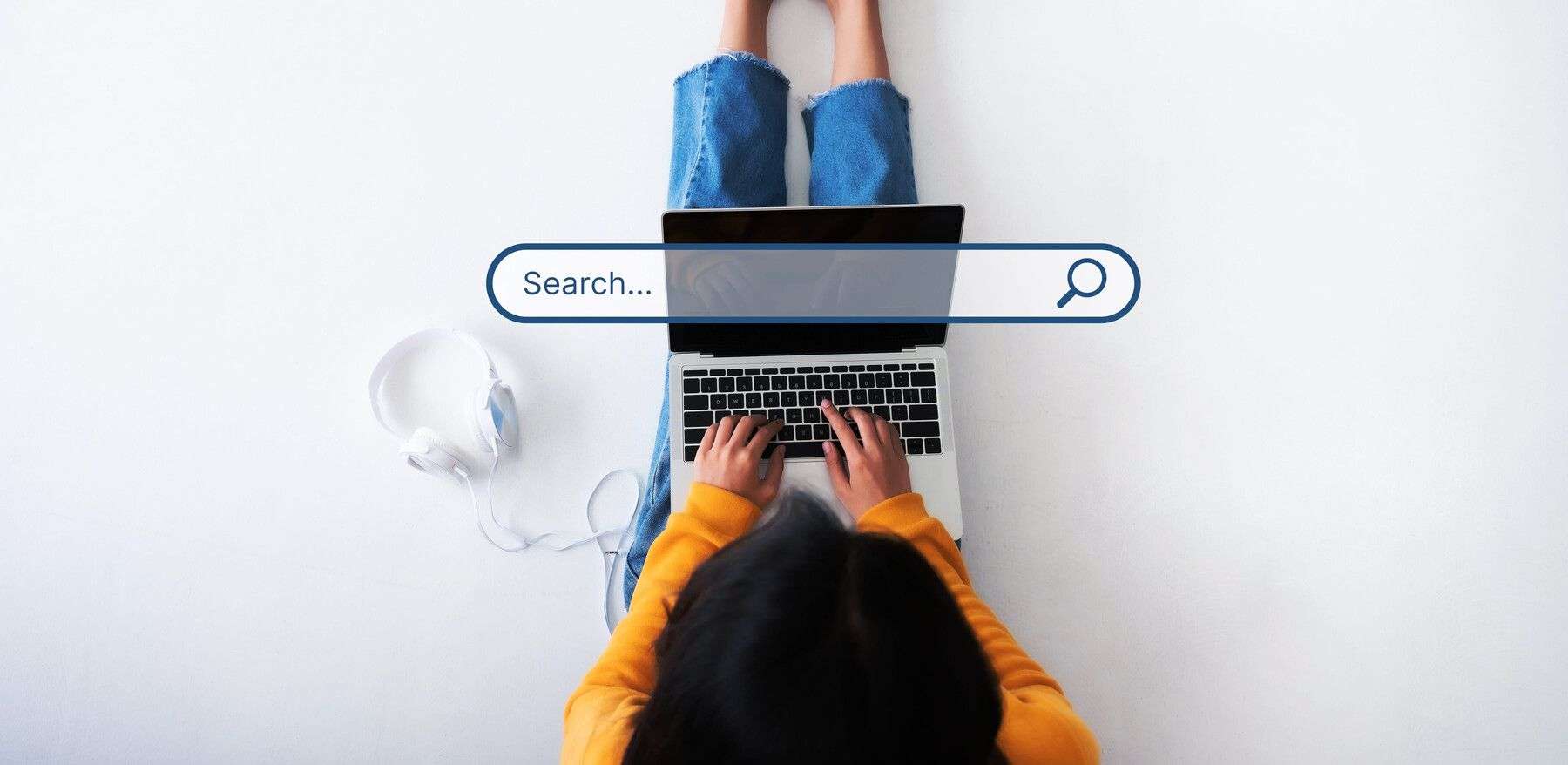 A person is seen from a top-down perspective, lying on their back on a pristine white floor or surface, wearing a bright yellow long-sleeved shirt and frayed blue denim jeans, with their bare legs slightly bent at the knees. Their dark hair is visible at the bottom of the frame as they focus on typing with both hands on the black keys of a silver laptop. Overlaid prominently across the laptop’s screen is a stylized, semi-transparent blue search bar graphic with the placeholder text “Search…” and a magnifying glass icon on the right. To the left of the laptop, a pair of white over-ear headphones with a white cable rests neatly on the white surface. The overall scene is clean, minimalist, and brightly lit, emphasizing the contrast between the warm yellow of the shirt, the cool blue of the jeans, and the stark white background, with no visible facial expression since the person’s head is turned away from the camera.